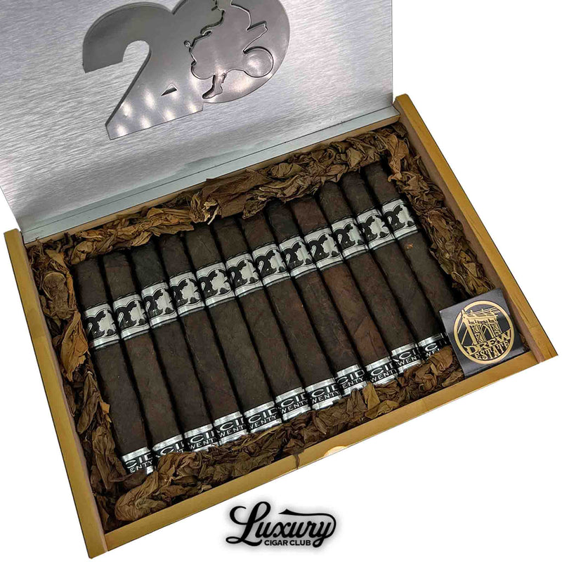 Close-up of Acid 20 Maduro Robusto cigars in open box with dark, box-pressed wrappers, silver and black bands, decorative tobacco leaf padding, a commemorative coin, and the Luxury Cigar Club logo below.