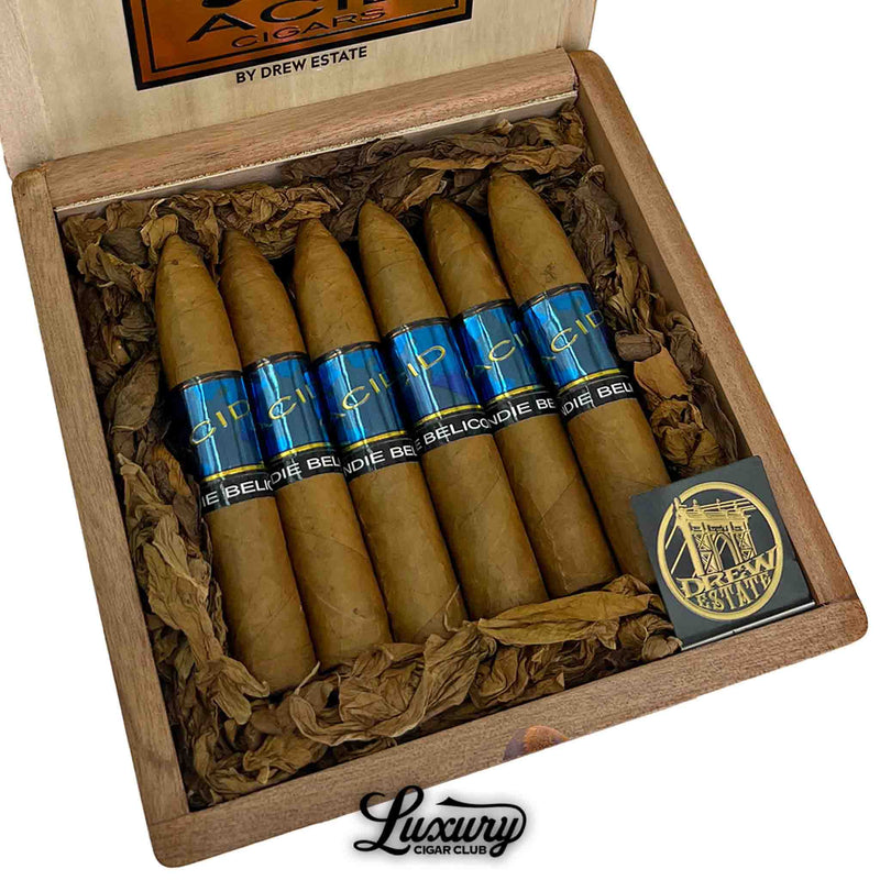 Close-up of Acid Blondie Belicoso cigars by Drew Estate, showcasing pointed heads, light Connecticut wrappers, and vibrant blue bands labeled “Blondie Belicoso.” Cigars are arranged in a cedar box with shredded tobacco and the Drew Estate logo.