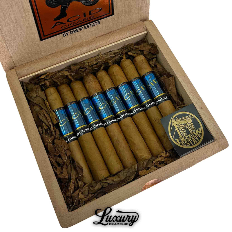 Close-up of Acid Blondie cigars by Drew Estate, featuring short cigars with light Connecticut wrappers and bold blue bands labeled “Blondie.” Arranged in a cedar box with shredded tobacco and a Drew Estate medallion.
