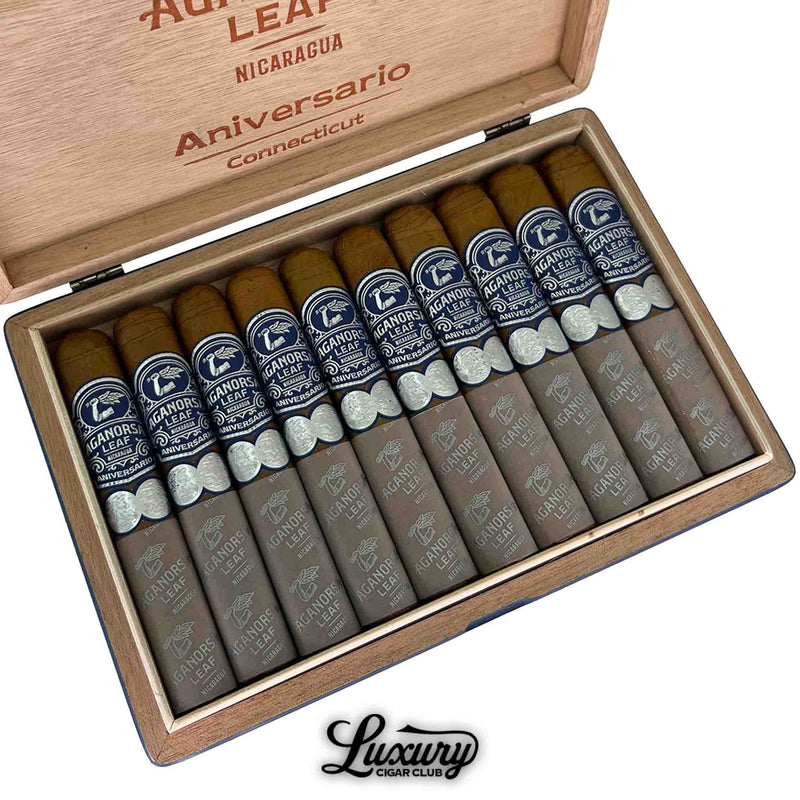 Close-up of Aganorsa Leaf Aniversario Connecticut Gran Robusto cigars in an open box, featuring ten cigars with golden Connecticut wrappers, dark blue and silver bands, and gray foot sleeves. The Aganorsa Leaf branding is prominently displayed on each cigar and on the inside lid.