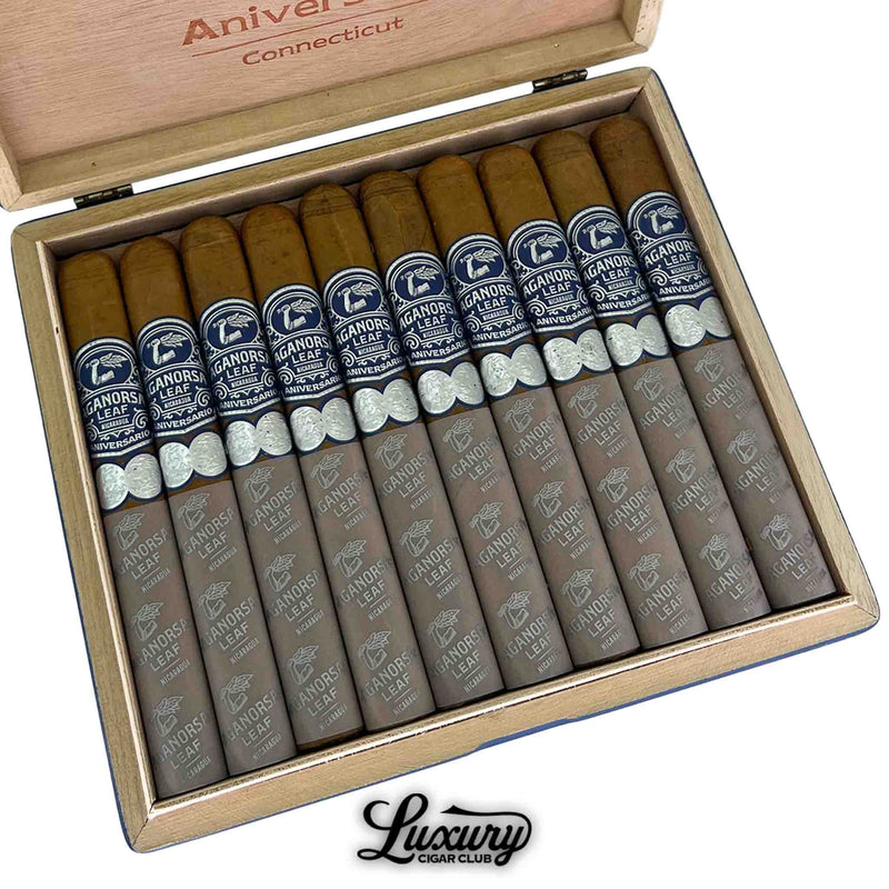 Close-up of an open box containing ten Aganorsa Leaf Aniversario Connecticut Toro cigars. Each cigar features a golden Connecticut wrapper with dark blue and silver bands, along with gray foot sleeves printed with the Aganorsa Leaf logo. The Luxury Cigar Club logo is shown beneath the box.