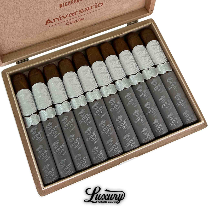 Close-up of an open box containing ten Aganorsa Leaf Aniversario Corojo Gran Toro cigars. Each cigar features a light brown wrapper with an ornate silver band and matching gray foot sleeve printed with the Aganorsa Leaf logo. The Luxury Cigar Club logo is visible below the box.