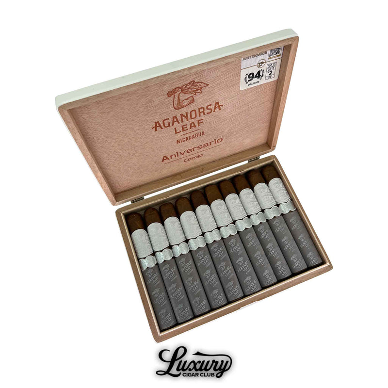 Open wooden box of Aganorsa Leaf Aniversario Corojo Gran Toro cigars, featuring ten cigars with silver bands and gray foot sleeves. The inside lid is branded with the Aganorsa Leaf logo, “Aniversario Corojo,” and a 94-point rating sticker. The Luxury Cigar Club logo appears below the box.