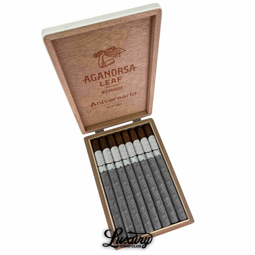 Open wooden box of Aganorsa Leaf Aniversario Corojo Lancero cigars, displaying ten slender cigars with silver bands and gray foot sleeves. The inside lid is engraved with the Aganorsa Leaf logo and “Aniversario Corojo Nicaragua” text. The Luxury Cigar Club logo appears below the box.