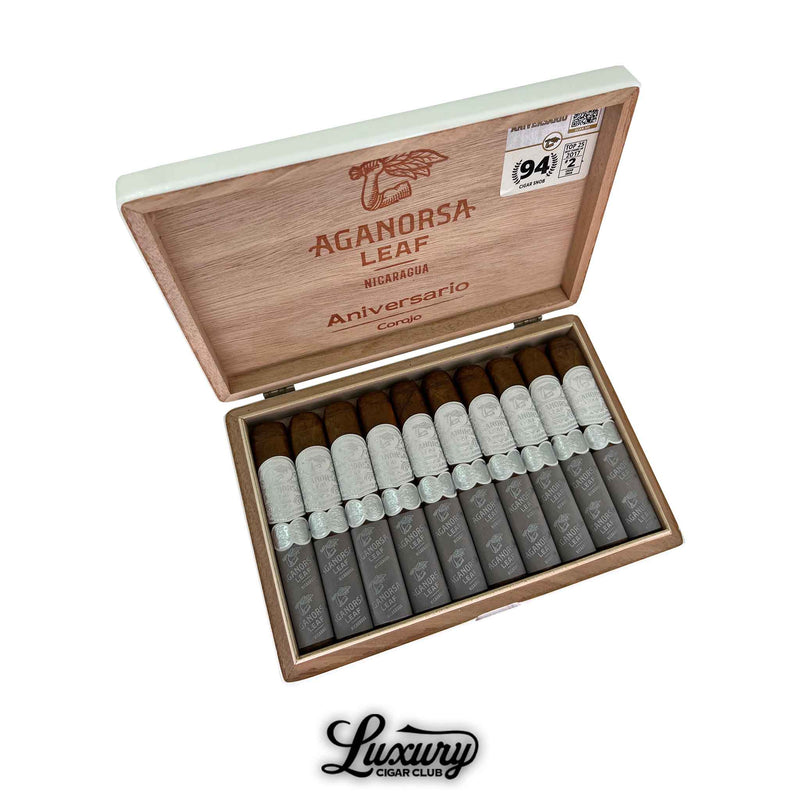 Open wooden box of Aganorsa Leaf Aniversario Corojo Robusto cigars, featuring ten cigars with silver bands and gray foot sleeves. The inside lid displays the Aganorsa Leaf logo, “Aniversario Corojo” branding, and a 94-rating sticker. The Luxury Cigar Club logo appears below the box.