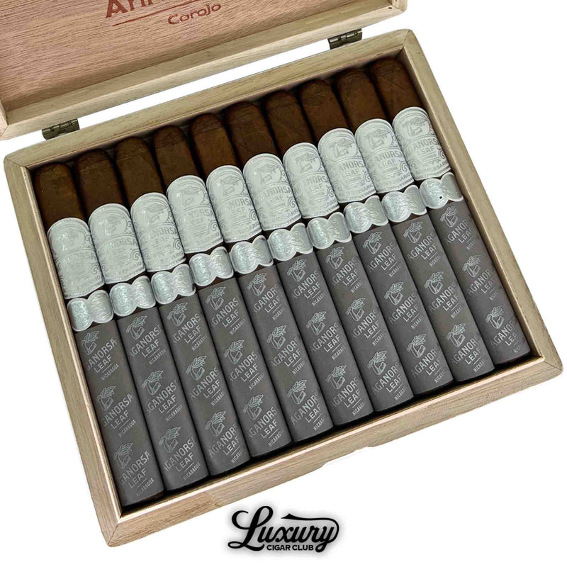 Close-up of an open box containing ten Aganorsa Leaf Aniversario Corojo Toro cigars. Each cigar features a light brown wrapper, a silver band, and a gray foot sleeve with the Aganorsa Leaf branding. The Luxury Cigar Club logo appears below the box.