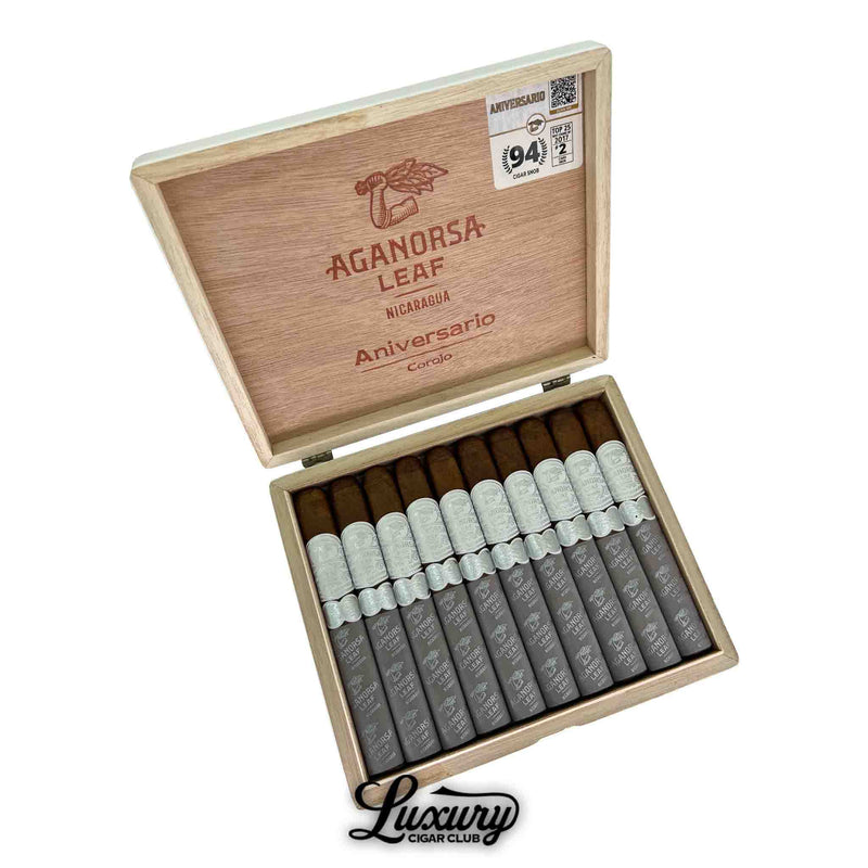 Open wooden box of Aganorsa Leaf Aniversario Corojo Toro cigars, featuring ten cigars with silver bands and gray foot sleeves. The inside lid displays the Aganorsa Leaf logo and “Aniversario Corojo” branding along with a “94 Rated” Cigar Aficionado sticker. The Luxury Cigar Club logo appears below the box.