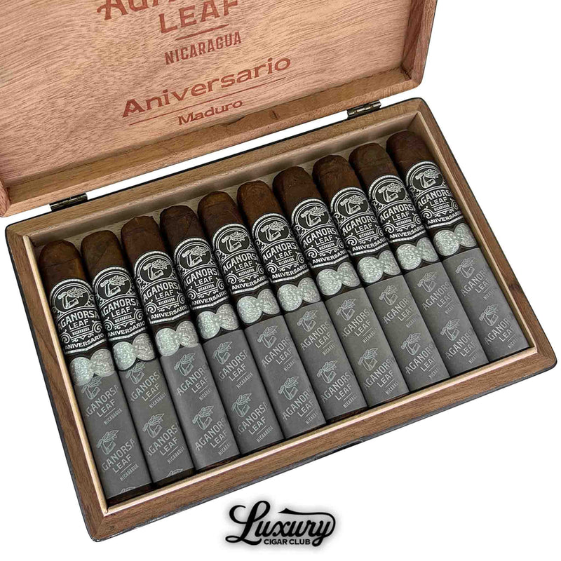 Close-up view of Aganorsa Leaf Aniversario Maduro Gran Robusto cigars in an open wooden box, showcasing dark maduro wrappers and elegant black-and-silver bands with decorative foot ribbons and crisp branding.
