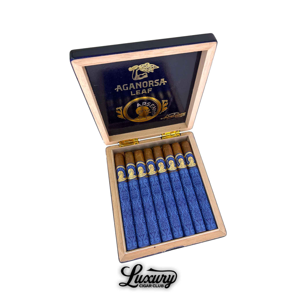 Open box of Aganorsa Leaf Arsenio Corona cigars with vivid blue and gold wrapping and golden-brown caps, arranged neatly under a glossy lid featuring gold Aganorsa Leaf and Arsenio branding. Luxury Cigar Club.