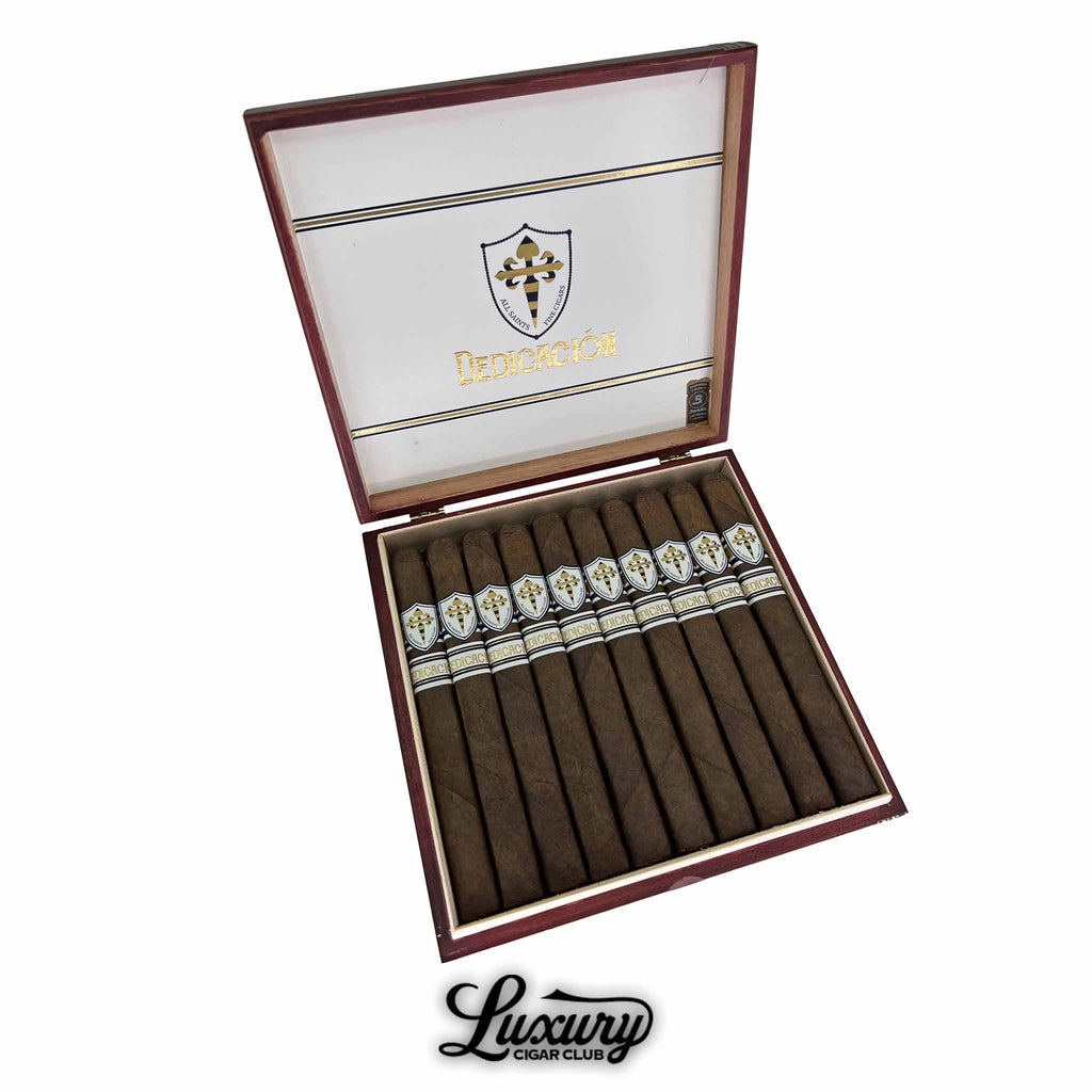Open box of All Saints Dedicación Coach cigars featuring twelve dark-wrapped cigars with elegant white and gold banding, presented by Luxury Cigar Club.