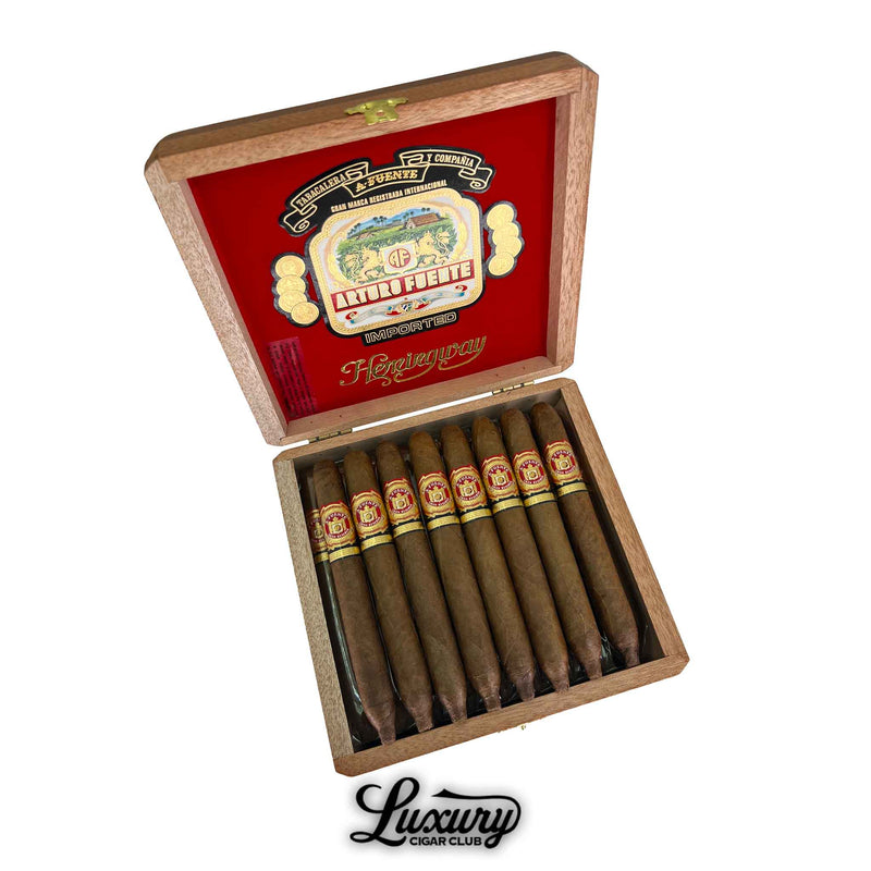 Open box of Arturo Fuente Hemingway Signature cigars displayed in a wooden case with a red velvet-lined lid featuring the Arturo Fuente logo, containing ten slender, torpedo-shaped cigars with red and gold bands, and a “Luxury Cigar Club” logo beneath the box.