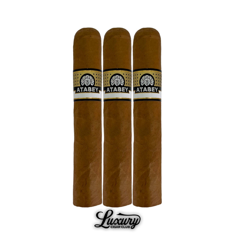 Three Atabey Brujos cigars in short robusto format, light brown wrapper, black and gold Atabey logo bands, and shiny gold decorative foot bands, presented upright with Luxury Cigar Club branding.