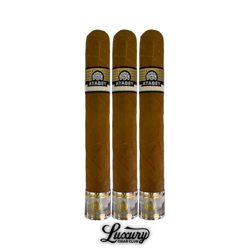 Three Atabey Misticos cigars in larger vitola, smooth Connecticut shade wrapper, black and gold Atabey bands, golden foil foot bands, presented upright with Luxury Cigar Club logo beneath.