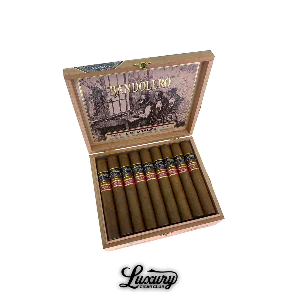 Open wooden box of Bandolero Serie C Colosales cigars, showcasing 25 large format sticks with golden brown wrappers and dual black and red bands, displayed by Luxury Cigar Club.