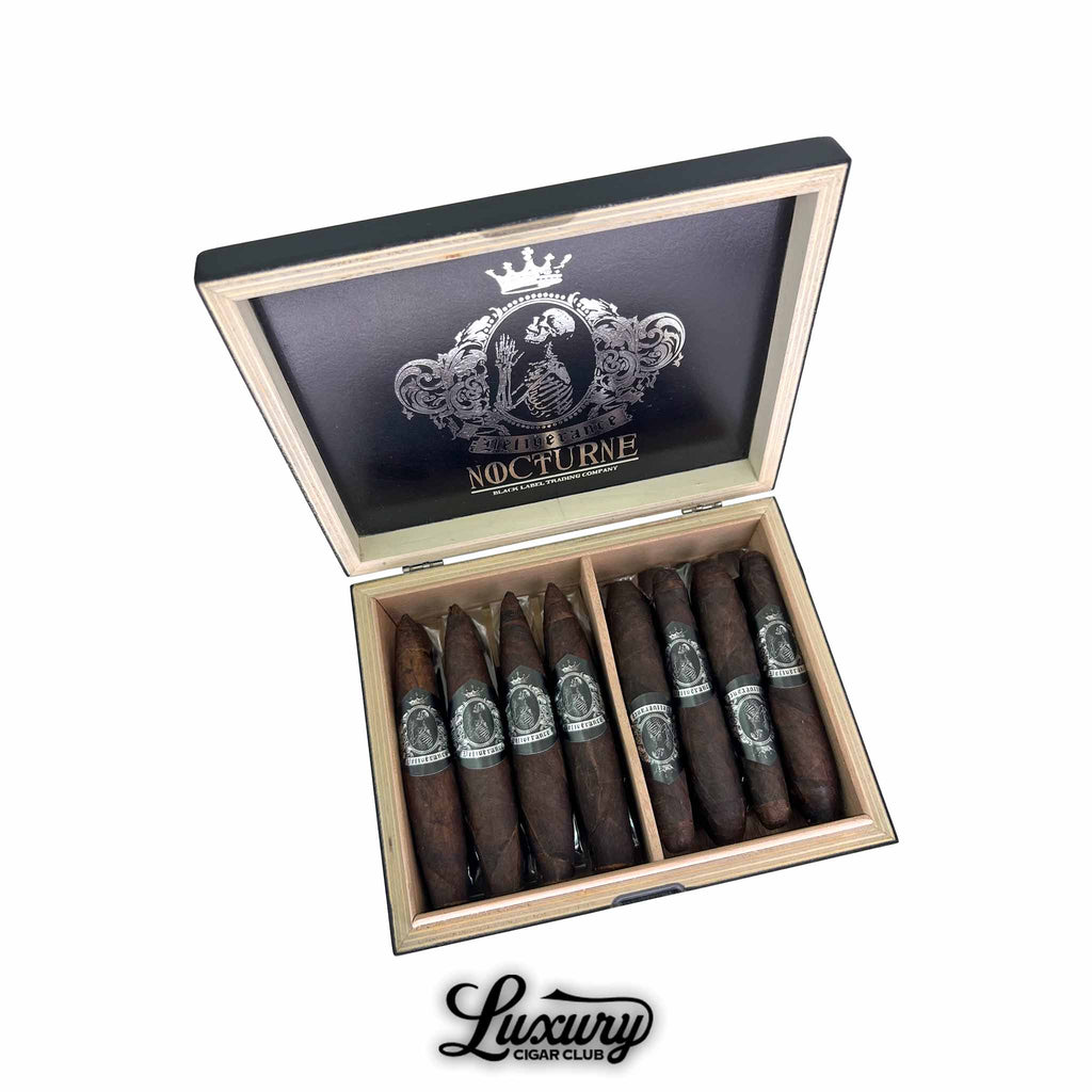 Luxury Cigar Club product photo showing an open Black Label Trading Co. Deliverance Nocturne Limited Edition combo box filled with dark Maduro cigars in two vitolas, each banded with an ornate silver skeleton crest, displayed inside a natural wood interior beneath a black lid with silver Nocturne branding.