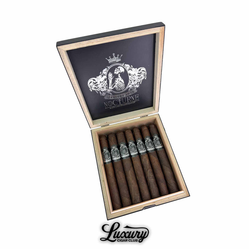 Open box of Black Label Trading Co. Deliverance Nocturne Toro cigars displayed in two rows with dark wrappers and silver skeletal bands, shown on a wood interior box lid featuring the Nocturne crest, photographed by Luxury Cigar Club.