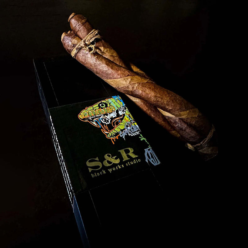 Black Works Studio S&R Limited Edition Lancero Culebra cigars entwined in a twisted trio presentation beside the signature snake artwork box, available at Luxury Cigar Club.