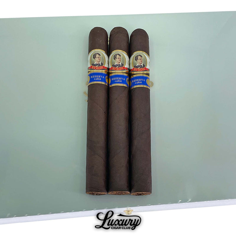Close-up of Byron 19th Century Epique Poemas cigars with dark wrappers and blue and gold Reserva 4 Años bands, resting on a white humidor lid at Luxury Cigar Club.