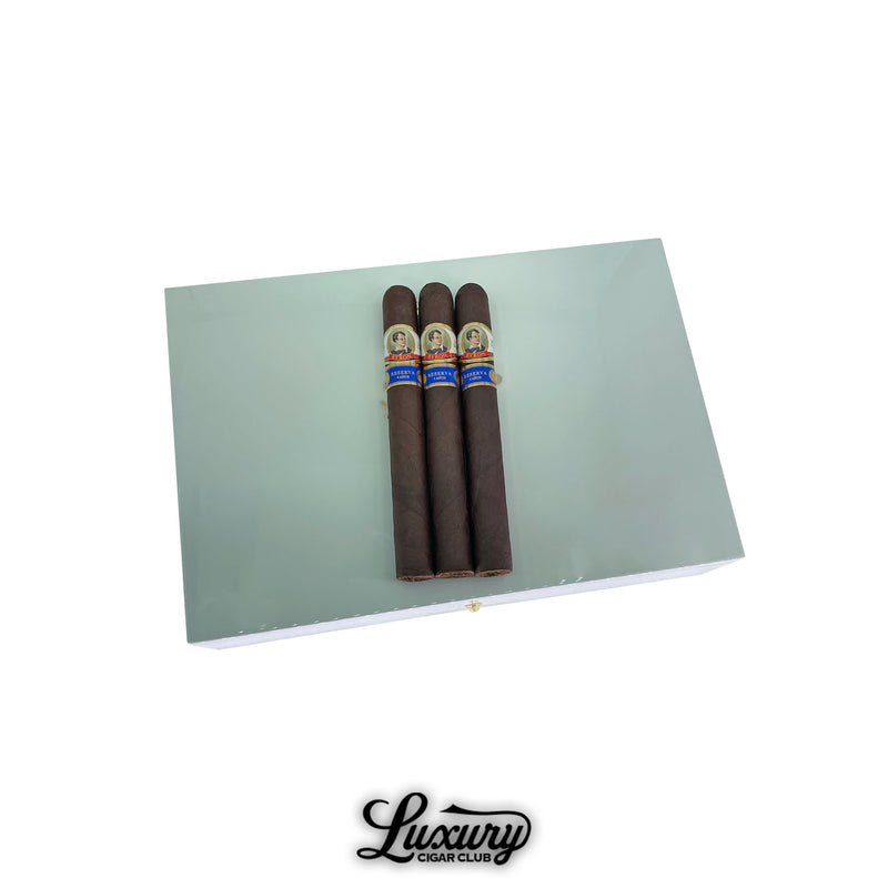 Byron 19th Century Epique Poemas cigars displayed on a white lacquered humidor box with three dark-wrapped cigars on top, available at Luxury Cigar Club.