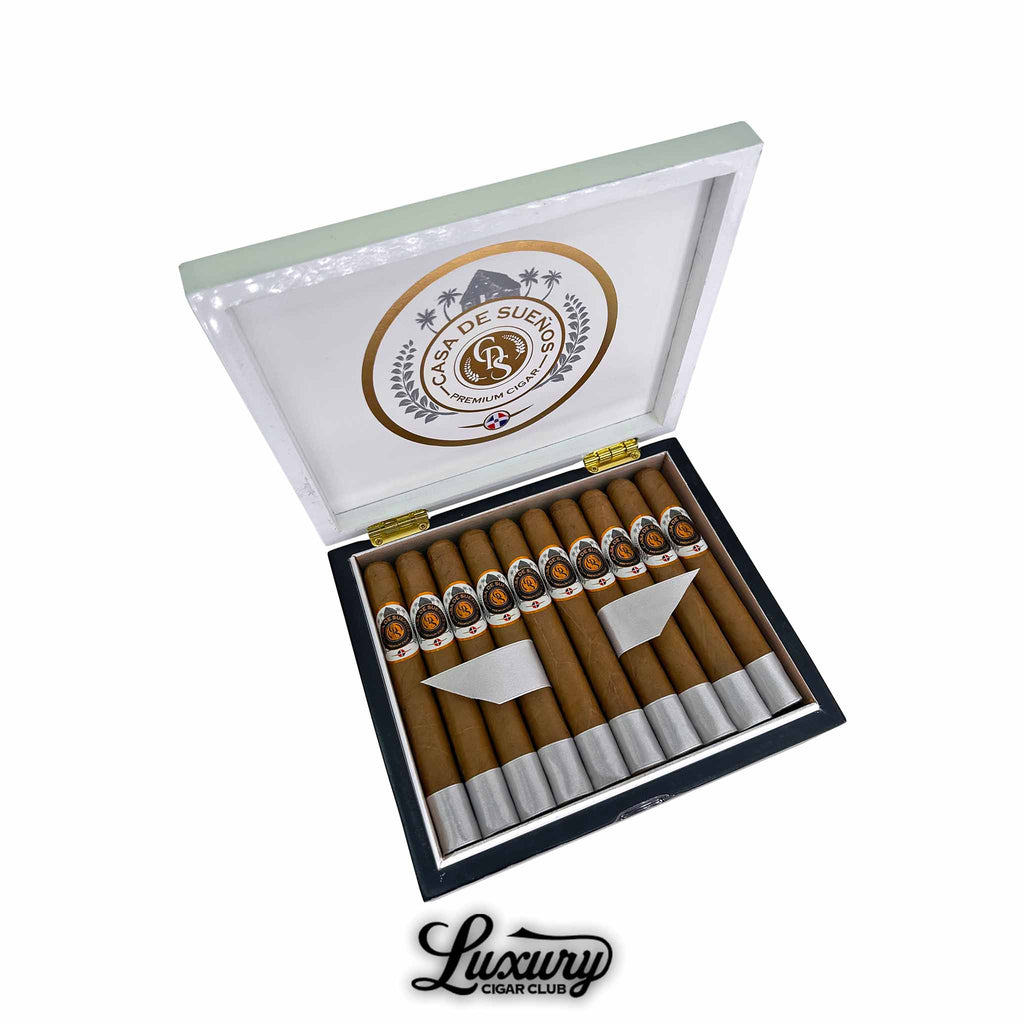Open box of Casa de Sueños Angelito Corona cigars with smooth golden wrappers and silver foot bands. The inside lid features the Casa de Sueños logo in gold and black. Luxury Cigar Club logo below.
