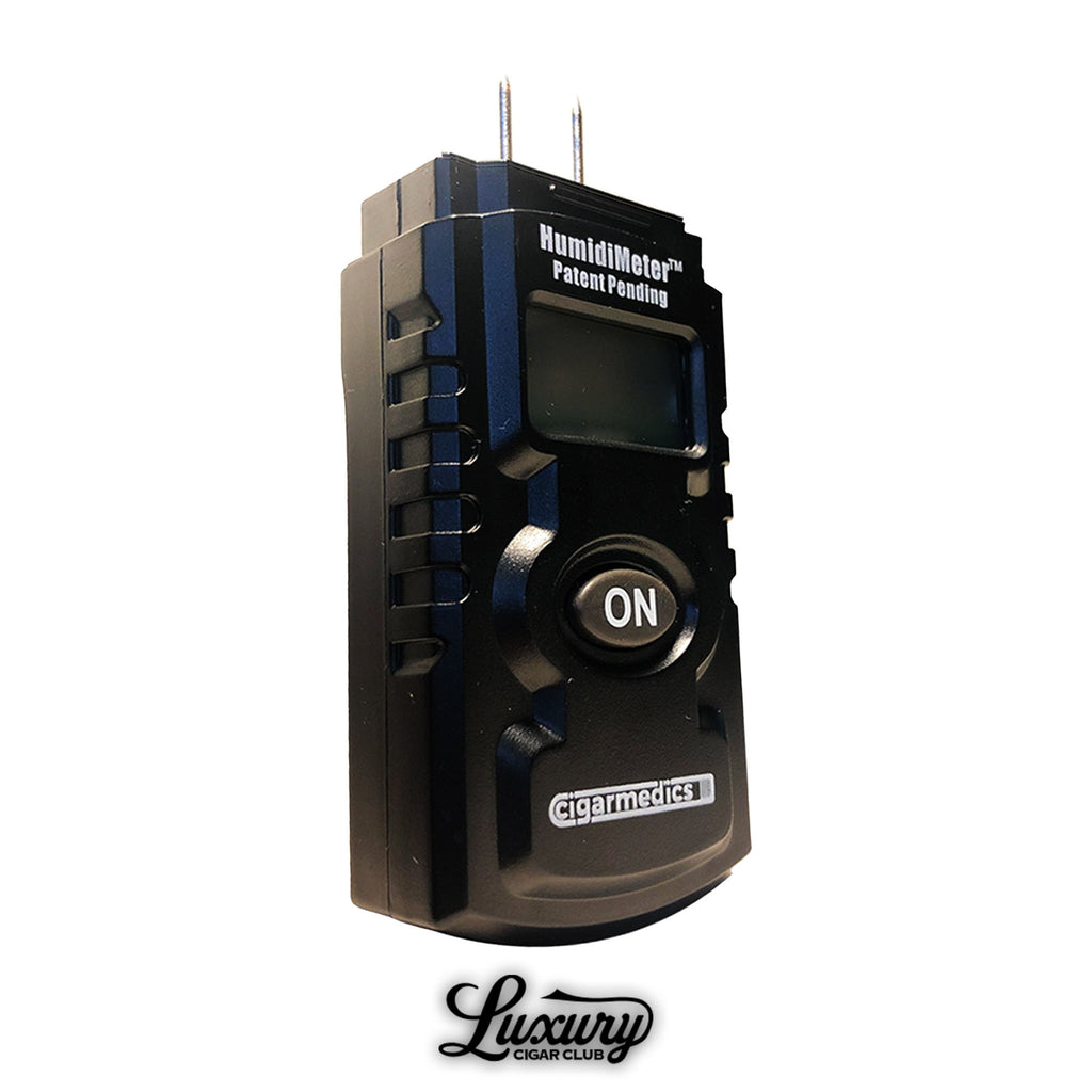 CigarMedics HumidiMeter digital cigar hygrometer with black rugged design, large ON button, dual metal probes for accurate moisture readings, and small display screen, shown upright with Luxury Cigar Club logo at the bottom.
