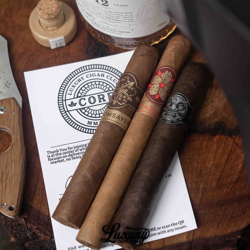 Three premium cigars from Luxury Cigar Club’s Core subscription, Doomsayer, Farce, and Big Payback Maduro cigar, arranged on top of a CORE membership card, placed beside a bottle of whiskey, a cork stopper, and a wooden-handled cigar cutter on a rustic wooden surface.