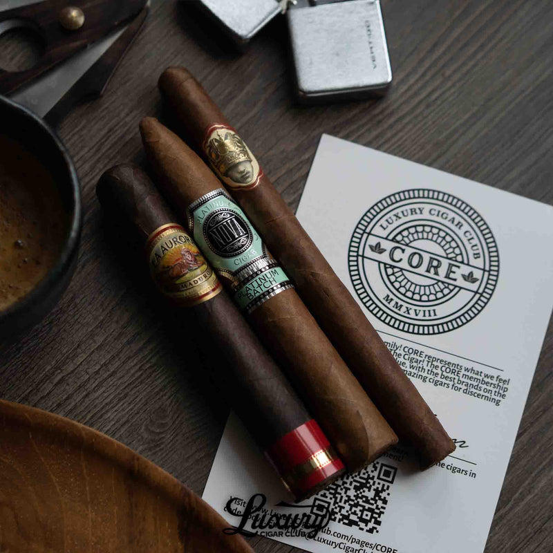 Three premium cigars from Luxury Cigar Club’s Core subscription—La Aurora Maduro, Nova Platinum Batch, and another banded cigar, resting on a CORE membership card beside a cup of coffee, cigar scissors, and a lighter on a wooden surface.