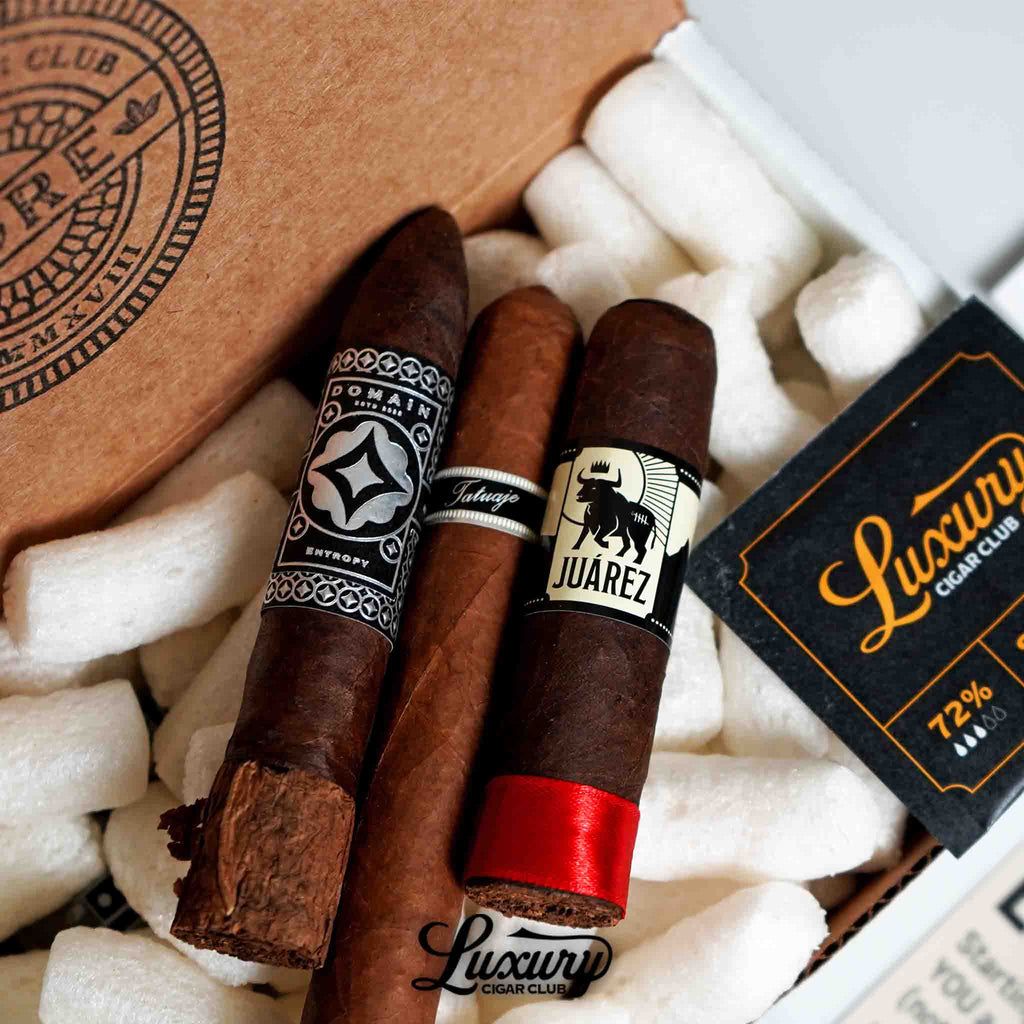 Close-up of a Luxury Cigar Club subscription box featuring three premium cigars, Domain Entropy, Tatuaje, and Juárez, resting on white packing material alongside a black and gold Luxury Cigar Club humidity pack.