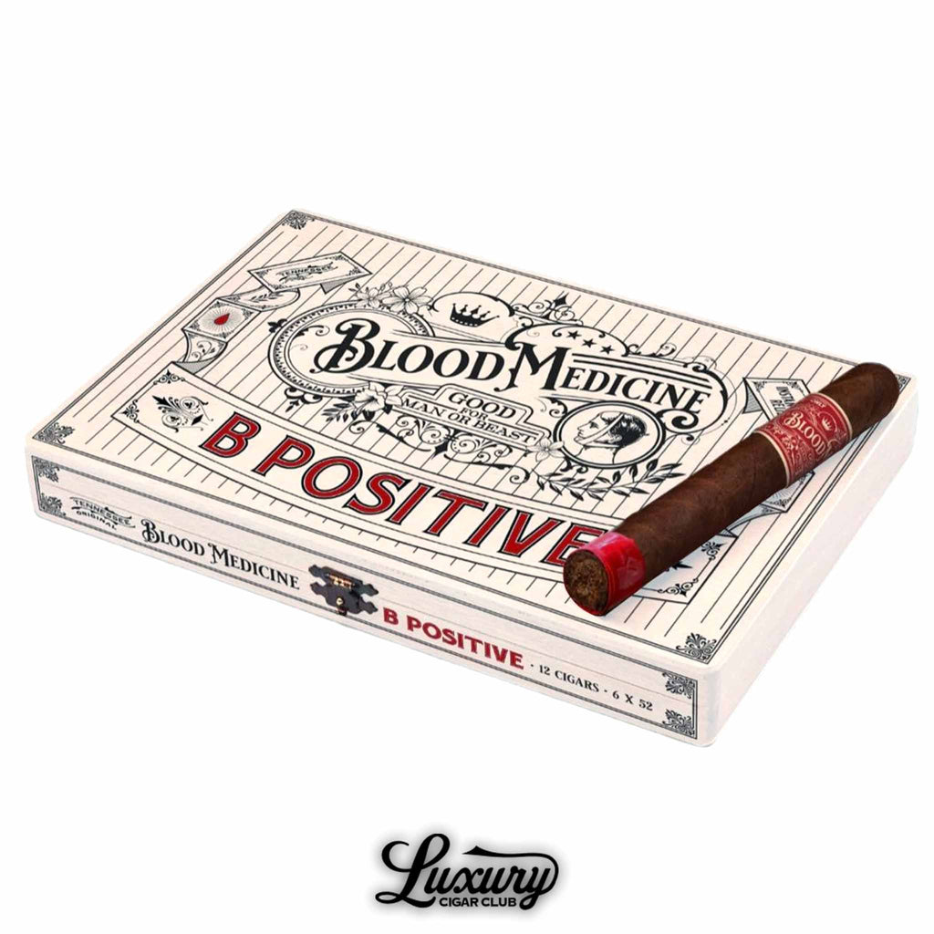 Crowned Heads Blood Medicine B Positive Limited Edition box of cigars with one cigar resting on top, featuring ornate vintage-style white packaging with bold red lettering, and the Luxury Cigar Club logo displayed beneath.