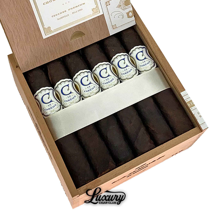 Close-up of Crowned Heads Le Carême cigars arranged in a wooden box, showcasing rich dark brown wrappers and detailed white-and-blue embossed bands. Luxury Cigar Club logo beneath.