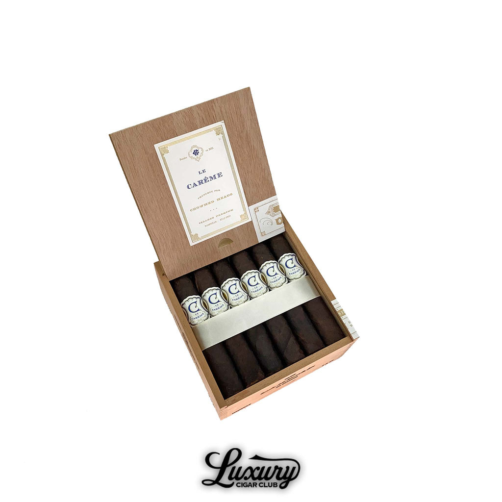 Open box of Crowned Heads Le Carême cigars with dark Maduro wrappers and elegant white and blue bands. The lid displays a cream and gold label design. Luxury Cigar Club logo below.