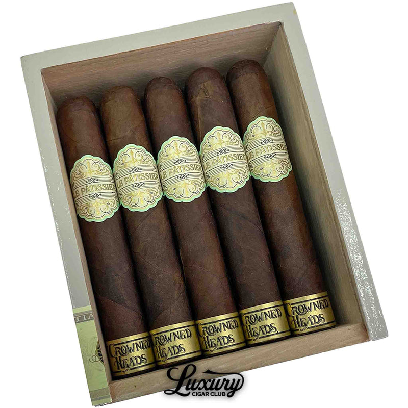Close-up of Crowned Heads Le Pâtissier No. 54 cigars showing smooth dark wrappers and intricate embossed gold and cream bands. Arranged in a light wooden box with Luxury Cigar Club logo beneath.