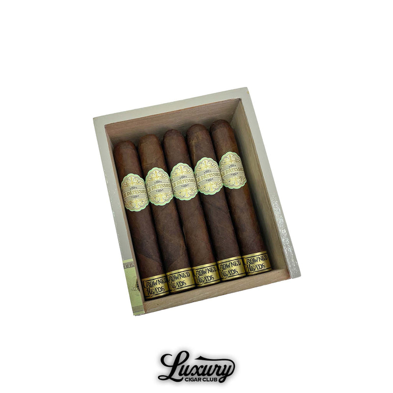Open box of Crowned Heads Le Pâtissier No. 54 cigars displayed in rows, each wrapped in a dark Maduro leaf with gold-trimmed labels. The presentation exudes a refined artisanal look. Luxury Cigar Club logo below.