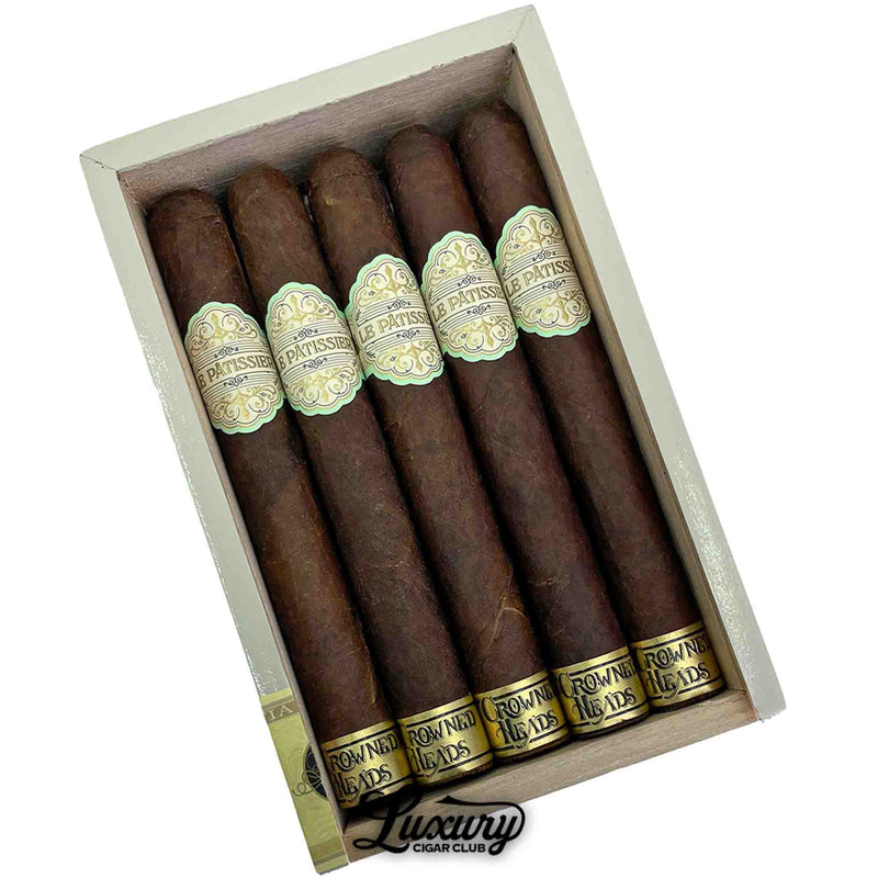 Close-up of Crowned Heads Le Pâtissier Senadores cigars highlighting their rich Maduro wrappers and elegant gold and cream labels. Cigars are displayed inside a light wood box. Luxury Cigar Club logo beneath.