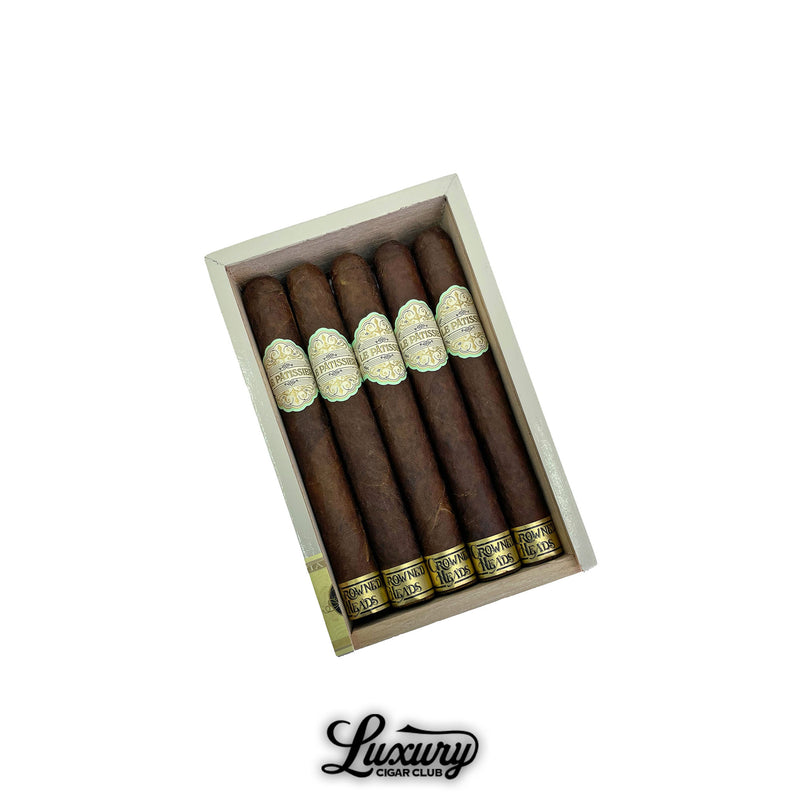 Open box of Crowned Heads Le Pâtissier Senadores cigars featuring dark chocolate-hued wrappers with ornate cream and gold bands. The cigars are neatly arranged inside a cream-colored box. Luxury Cigar Club logo below.