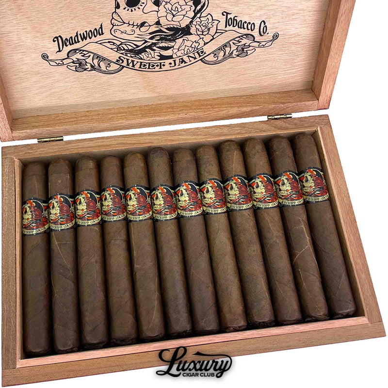 Close-up of Deadwood Sweet Jane Corona cigars arranged neatly in a wooden box, showcasing dark Maduro wrappers and detailed skull artwork on each band. Luxury Cigar Club logo beneath.