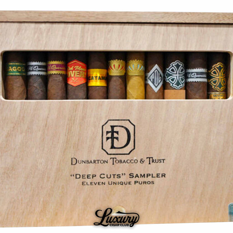 Close-up view of the Dunbarton Tobacco & Trust Deep Cuts Sampler showing eleven distinct cigars with detailed bands including Umbagog, Mi Querida, Triqui Traca, Muestra de Saka, Sin Compromiso, and other blends, highlighting wrapper color and branding inside the wooden sampler box for Luxury Cigar Club.