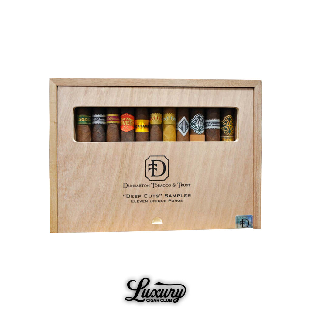 Dunbarton Tobacco & Trust Deep Cuts Sampler box featuring eleven unique puros displayed behind a clear window in a natural wood slide-lid box, showcasing premium cigars from Umbagog, Mi Querida, Triqui Traca, Muestra de Saka, Sin Compromiso, and more, photographed for Luxury Cigar Club.