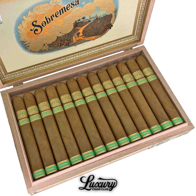 Close-up view of Dunbarton Tobacco & Trust Sobremesa Brûlée Blue Wagashi Toro cigars inside an open wooden box, revealing the smooth golden-brown Connecticut-shade wrappers and detailed green and gold Sobremesa bands. The ornate lid artwork emphasizes the cigar’s artisanal quality and refined construction. Luxury Cigar Club.