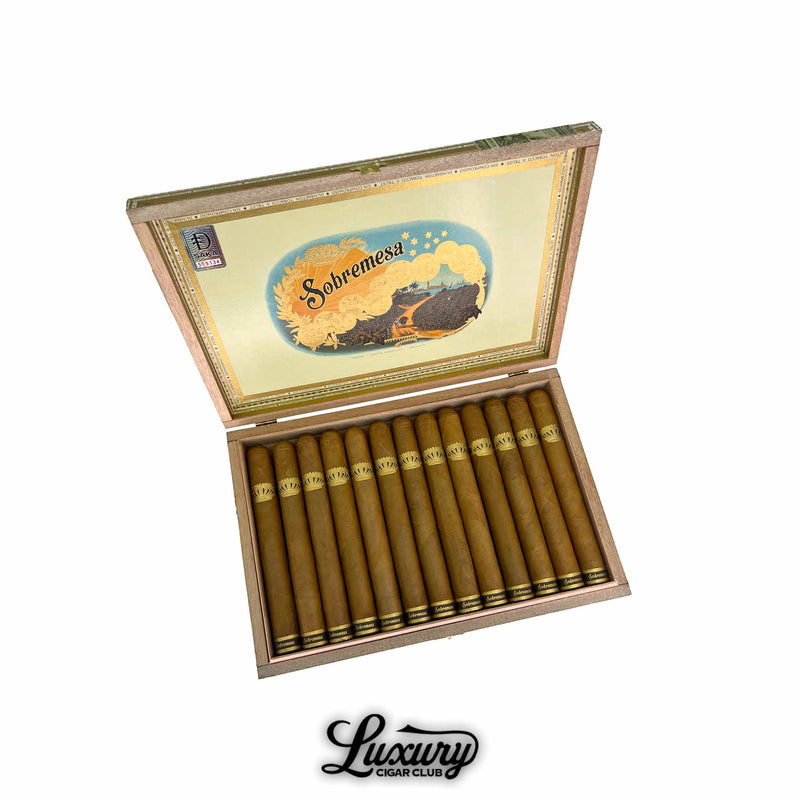 Open box of Dunbarton Tobacco & Trust Sobremesa Brûlée Double Corona cigars featuring long, golden Connecticut wrappers and detailed gold crown bands, shown under classic Sobremesa lid artwork. Luxury Cigar Club logo beneath.