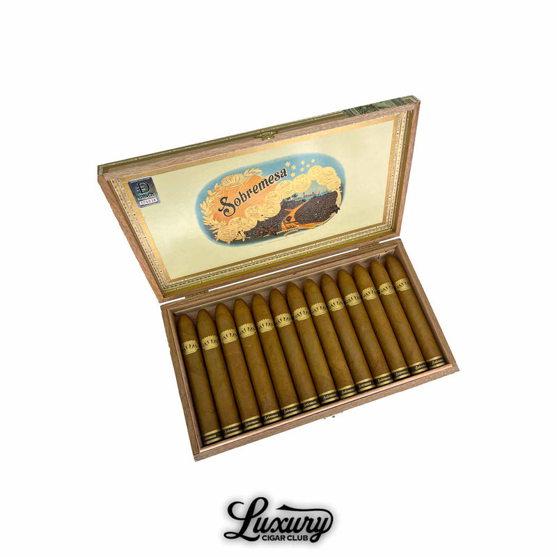 Open wooden box of Dunbarton Tobacco & Trust Sobremesa Brûlée Gordo cigars featuring light Connecticut Shade wrappers and elegant gold crown bands beneath vibrant Sobremesa artwork on the inside lid. Luxury Cigar Club logo below.