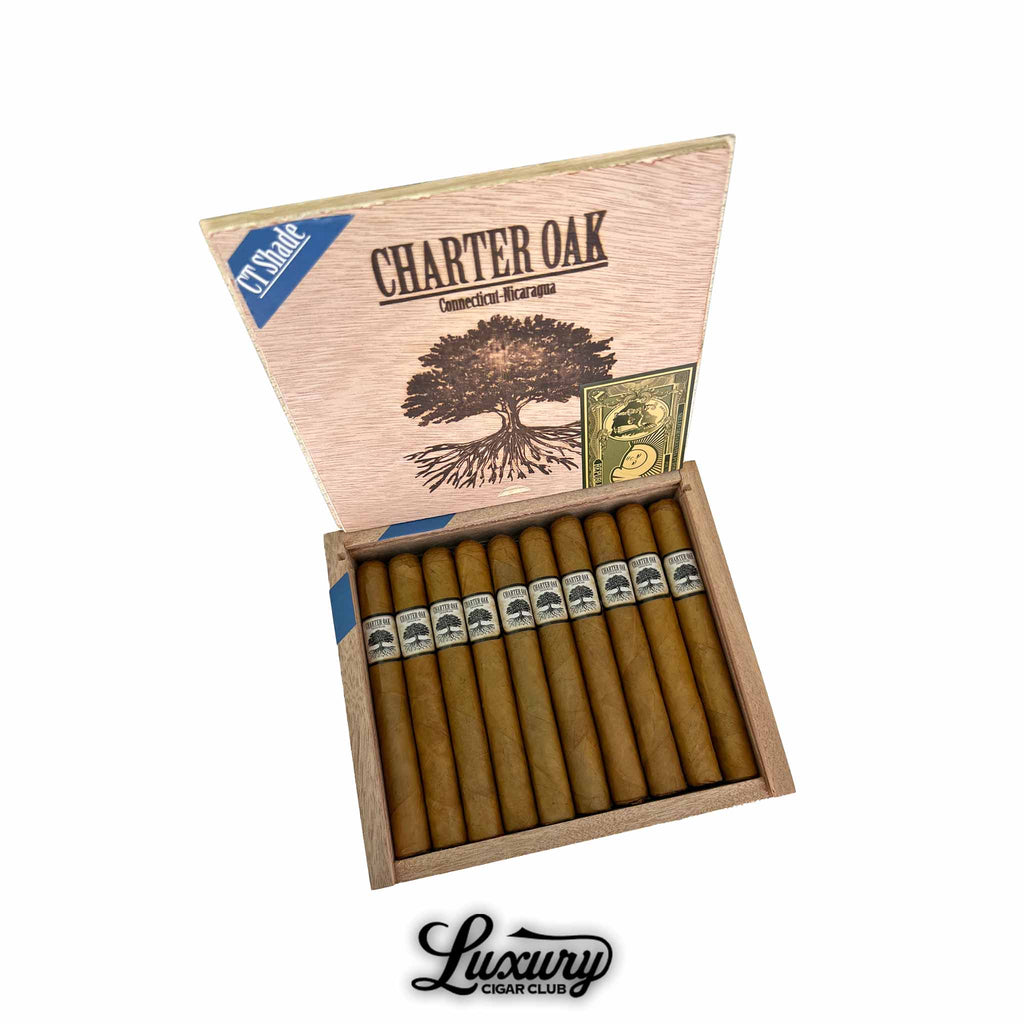 Open wooden box of Foundation Charter Oak Connecticut Petite Corona cigars featuring light golden-brown wrappers and classic black-and-white tree logo bands. Lid displays “Charter Oak Connecticut Nicaragua” branding. Luxury Cigar Club logo below.