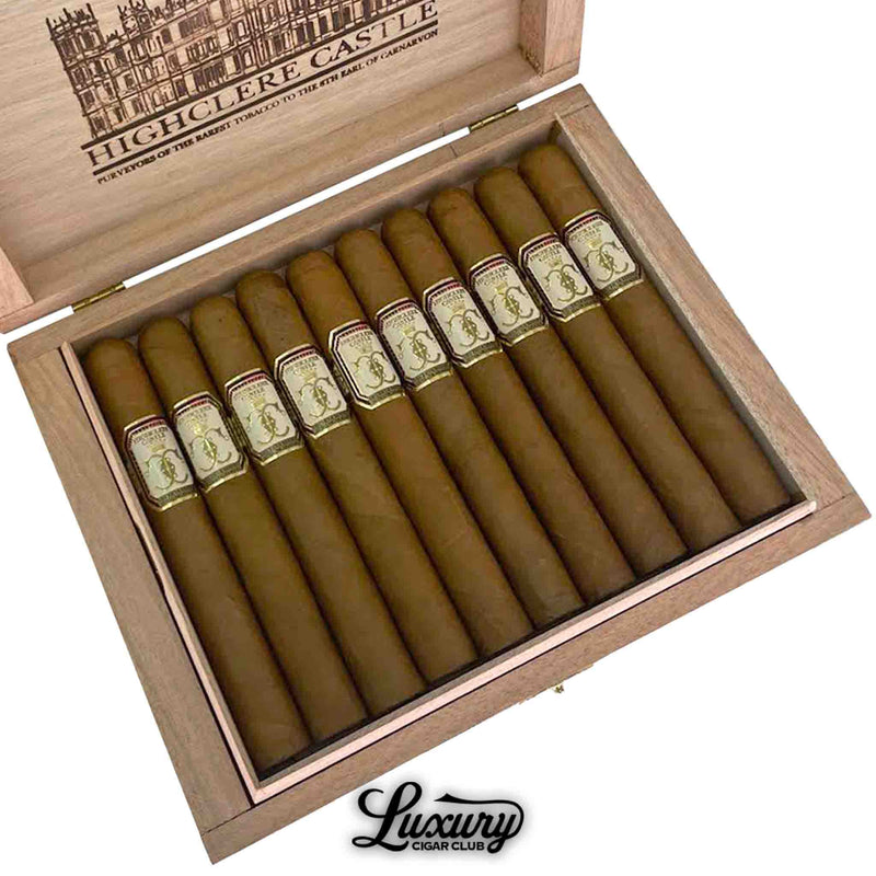 Close-up of an open box of Foundation Highclere Castle Edwardian Corona cigars, showing 10 smooth, light brown cigars with ornate cream, gold, and red bands. The wooden lid features an intricate illustration of Highclere Castle with branded text, and “Luxury Cigar Club” logo is visible below the box.