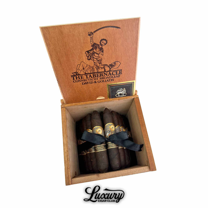 Open wooden box of Foundation The Tabernacle Broadleaf Goliath Limited Edition cigars showing rich dark wrappers tied with a black ribbon, with artwork of David and Goliath on the inside lid. Luxury Cigar Club logo beneath.