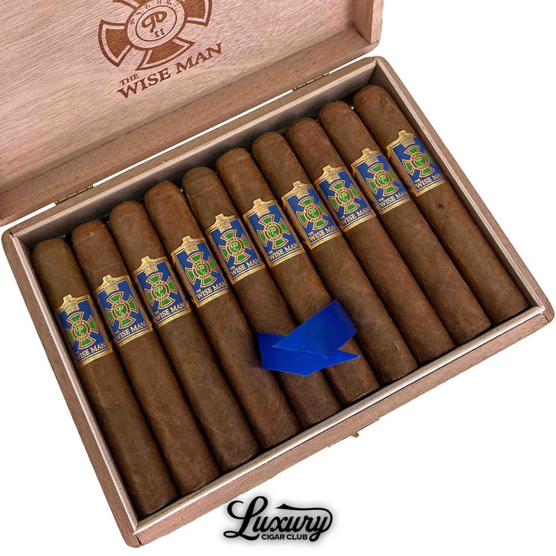 Close-up of Foundation The Wise Man Corojo Corona cigars in an open wooden box from Luxury Cigar Club. Each cigar features a golden brown wrapper with a detailed blue, green, and gold band. A blue ribbon lies across the center for elegant presentation, with “The Wise Man” branding engraved inside the lid.