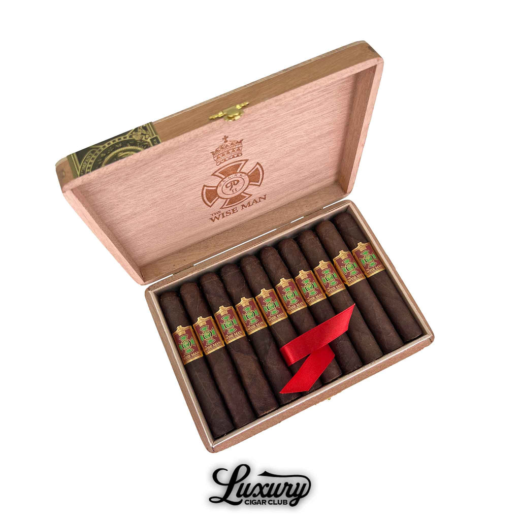 Open box of Foundation The Wise Man Maduro Corona cigars from Luxury Cigar Club. The cigars feature a dark, oily wrapper and are banded with vibrant red and green labels showcasing traditional artwork. A red ribbon secures the cigars in a natural wood box, with the Wise Man logo and emblem engraved on the inside of the lid.