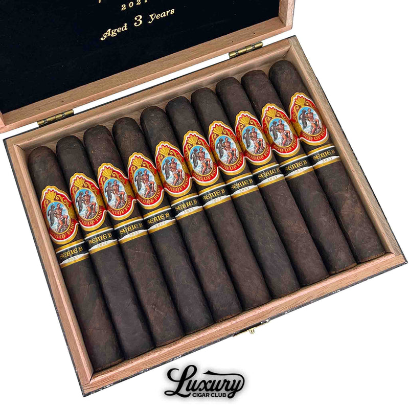 Close-up view of God of Fire Serie B Double Robusto cigars from 2021, aged 3 years, showcasing dark wrappers and ornate red, gold, and black bands with classical artwork, neatly arranged in a luxury wooden box with black velvet interior.