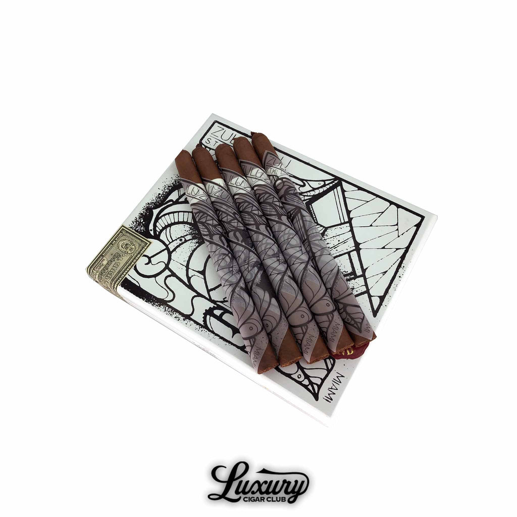 Five Gran Habano Zulu Zulu Mas Paz cigars displayed on top of a white, black-illustrated box featuring abstract artwork. Each cigar is wrapped in a striking, art-inspired sleeve with bold geometric designs, highlighting the collaboration with artist Mas Paz. The cigars feature dark brown wrappers and a refined tapered foot. The Luxury Cigar Club logo appears below. Luxury Cigar Club.
