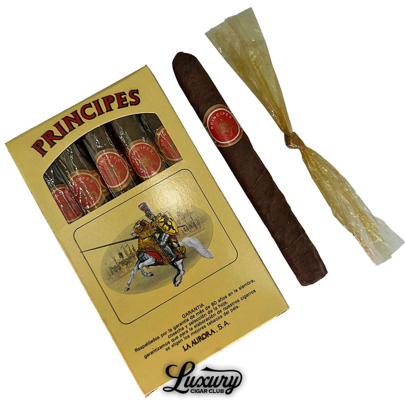 Close-up image of La Aurora Principes 5-count cigar box with six cigars displayed, one unwrapped and one in twisted cellophane. The cigars feature red and gold bands, and the box highlights a knight-on-horseback illustration, emphasizing the brand’s Dominican heritage. Ideal for showcasing cigars known for consistent flavor and value. 
