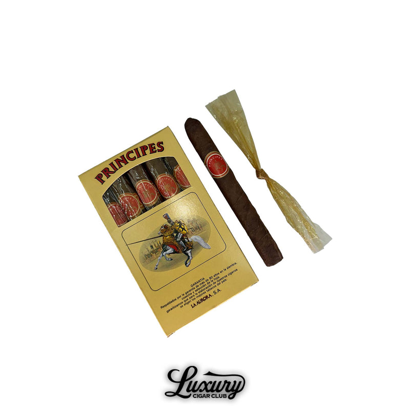 Box of La Aurora Principes cigars with six visible sticks, one unwrapped cigar, and one in twisted cello placed beside the box. The packaging showcases a red and gold banded design and a vintage knight illustration, emphasizing traditional Dominican craftsmanship. Ideal for showcasing budget-friendly, aged cigars.